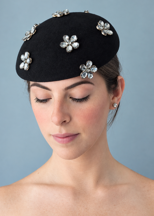 Model wearing the Émillie Noir percher hat adorned with five silver rhinestone flower brooches, photographed against a solid light blue-gray background.