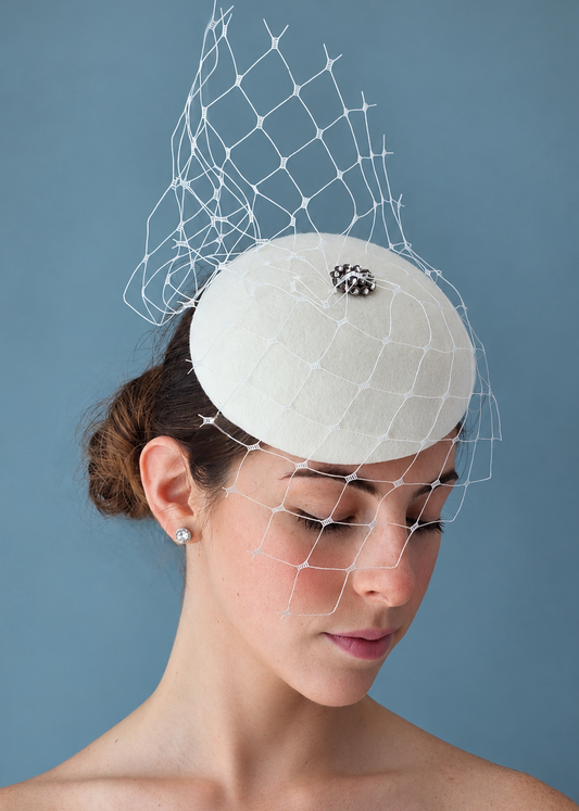 Model wearing the Lola percher hat with a white net veil and a silver jeweled accent on top, photographed against a muted blue-gray studio background.