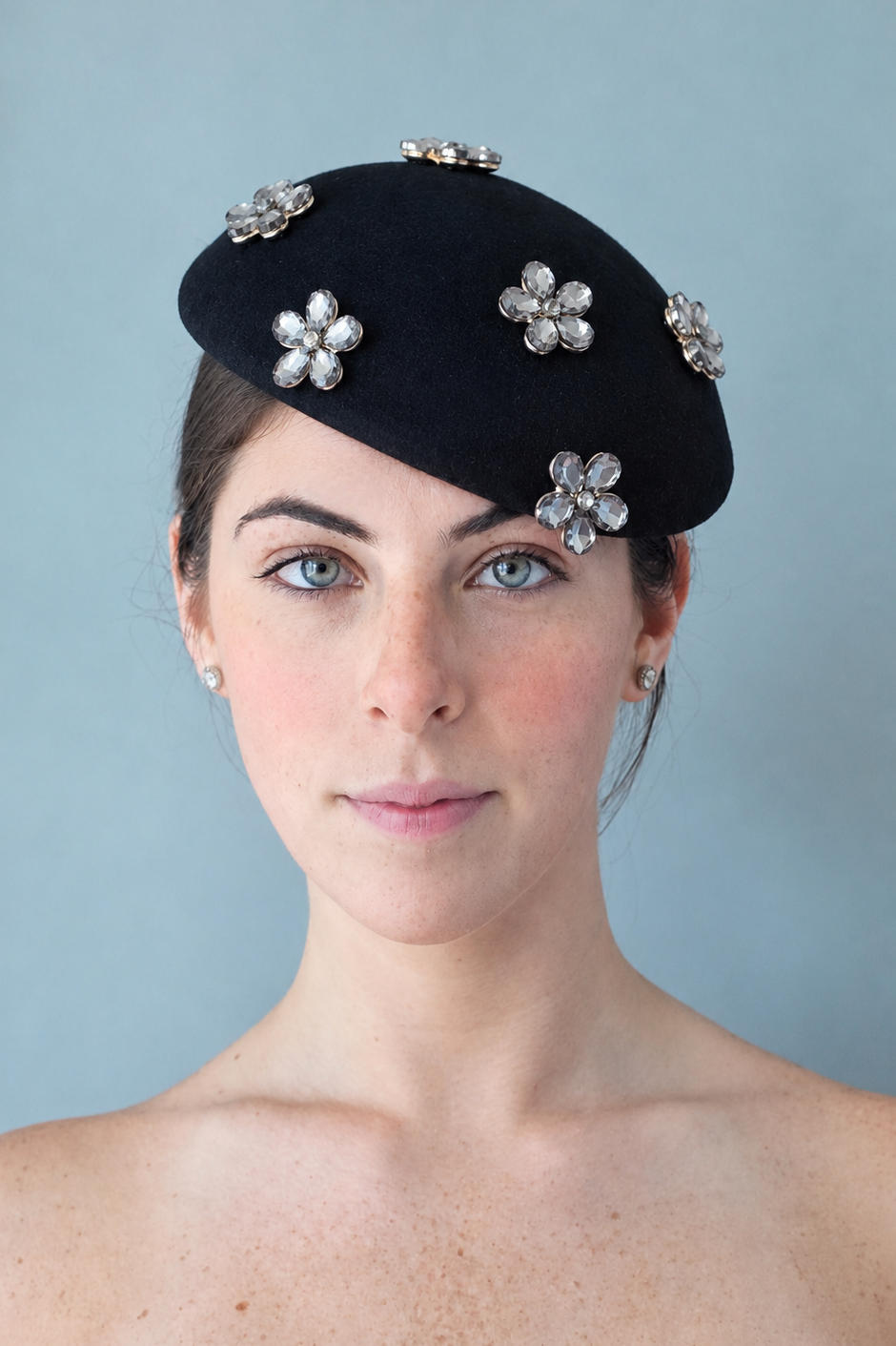 Model wearing the Émillie Noir percher hat adorned with five silver rhinestone flower brooches, photographed against a solid light blue-gray background.