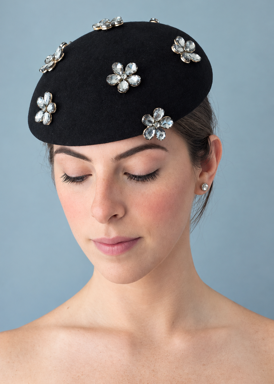Model wearing the Émillie Noir percher hat adorned with five silver rhinestone flower brooches, photographed against a solid light blue-gray background.