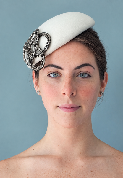 Model wearing the Fleur Percher Hat with a silver metallic knot embellishment, photographed against a solid blue-gray studio background.