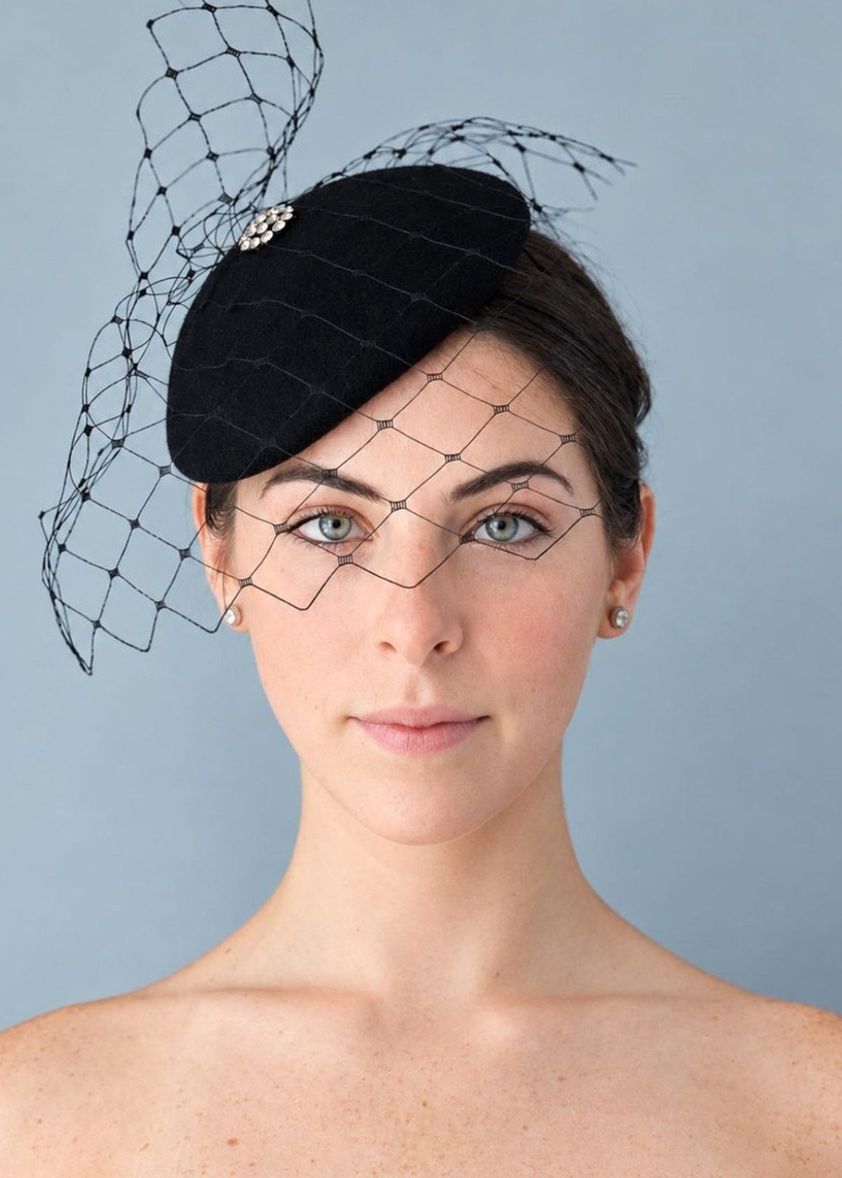 Model wearing the black Lola percher hat with a black birdcage veil and a small jeweled decorative accent, photographed against a plain light blue-gray studio background.