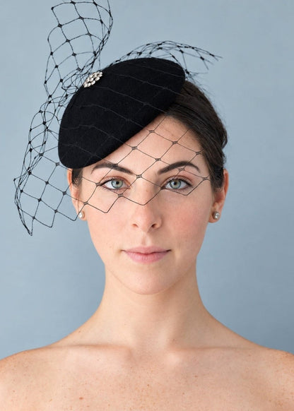 Model wearing the black Lola percher hat with a black birdcage veil and a small jeweled decorative accent, photographed against a plain light blue-gray studio background.