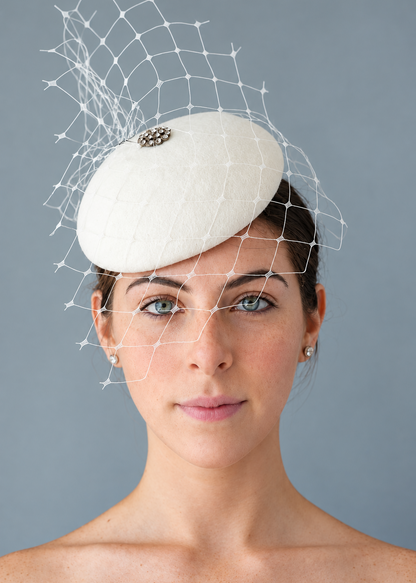 Model wearing the white Lola percher hat with a white net veil and a small decorative brooch, photographed against a plain gray studio background.