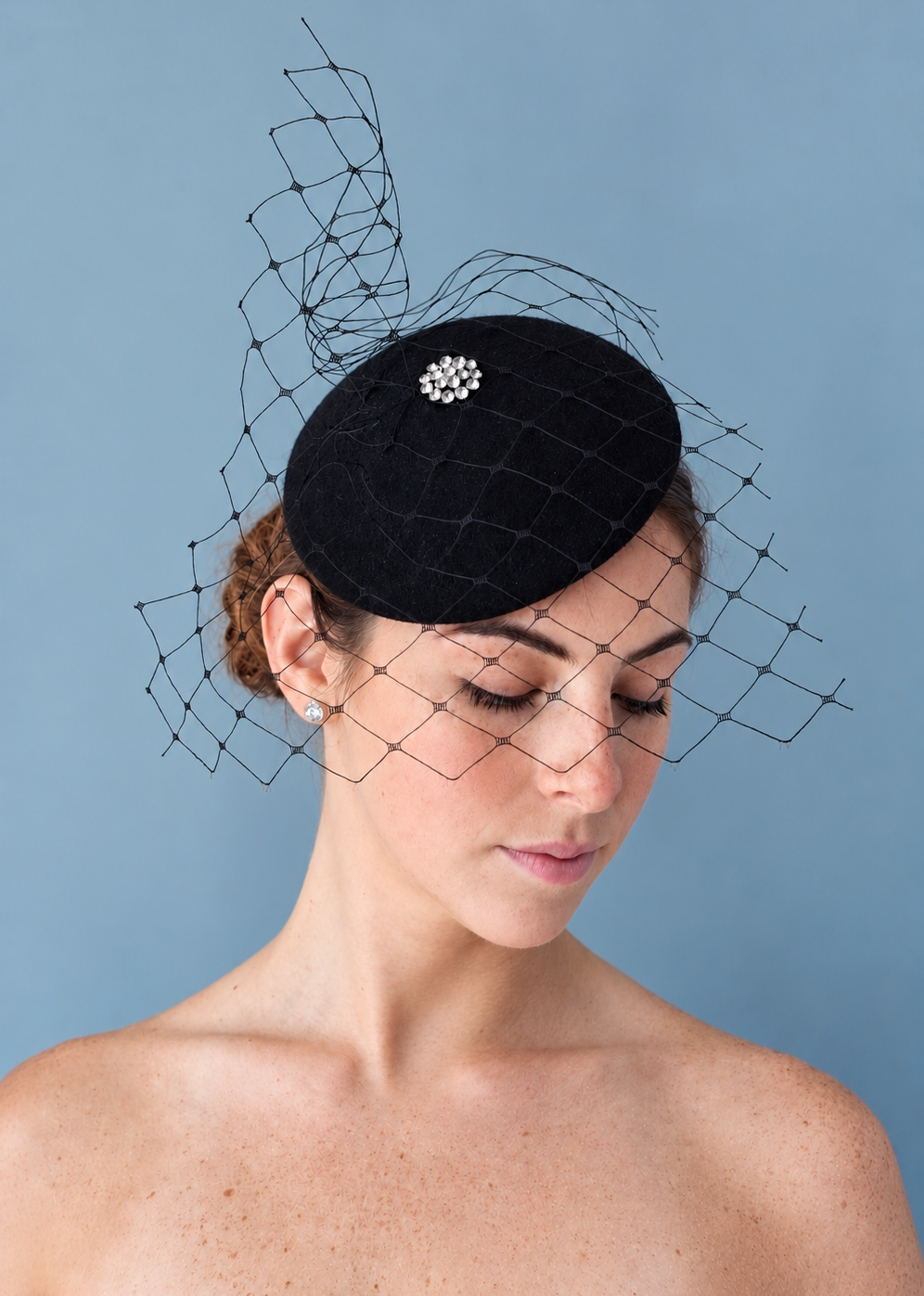 Model wearing the black Lola percher hat with a delicate black net veil and a silver crystal flower embellishment, photographed against a solid blue background.