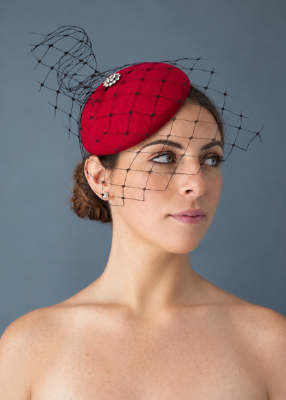 Model wearing the bred Lola percher hat with a delicate black net veil and a silver crystal flower embellishment, photographed against a solid blue background.