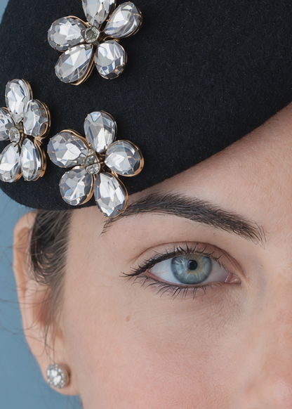 Close-up of the mini Emillie black percher hat adorned with three gold-tone flower embellishments set with faceted rhinestones.