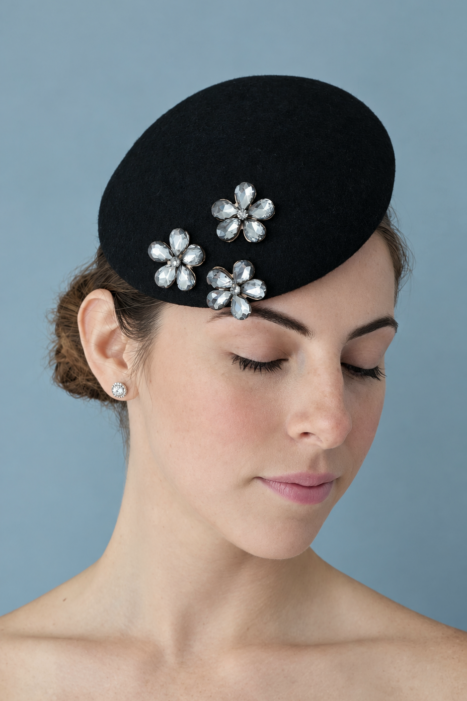  Mini Émilie Noir percher hat adorned with three rhinestone flower embellishments, set against a solid blue-gray background.