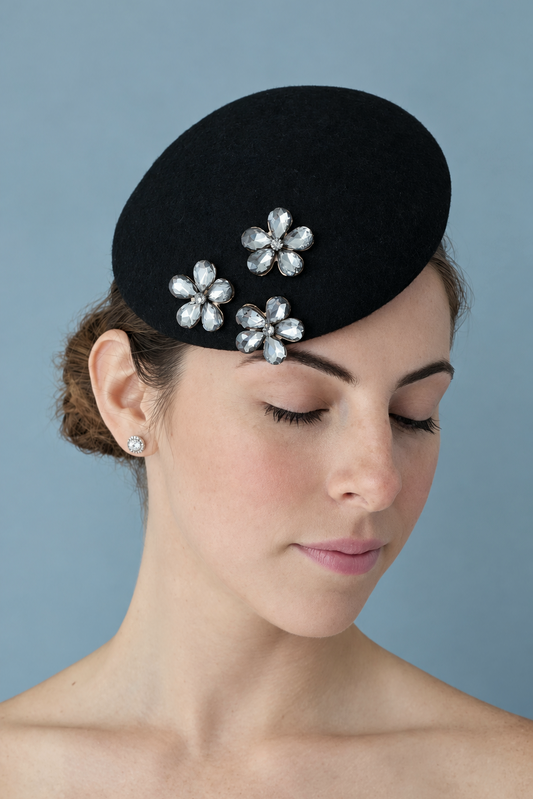  Mini Émilie Noir percher hat adorned with three rhinestone flower embellishments, set against a solid blue-gray background.