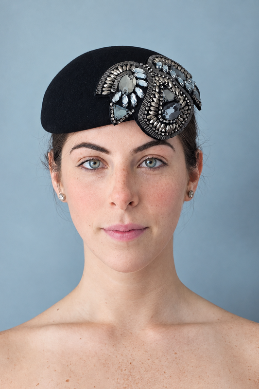 Simone  Percher Hat Front view. Black felt hat adorned with large, ornate silver and white rhinestone brooches, set against a solid blue-gray background.