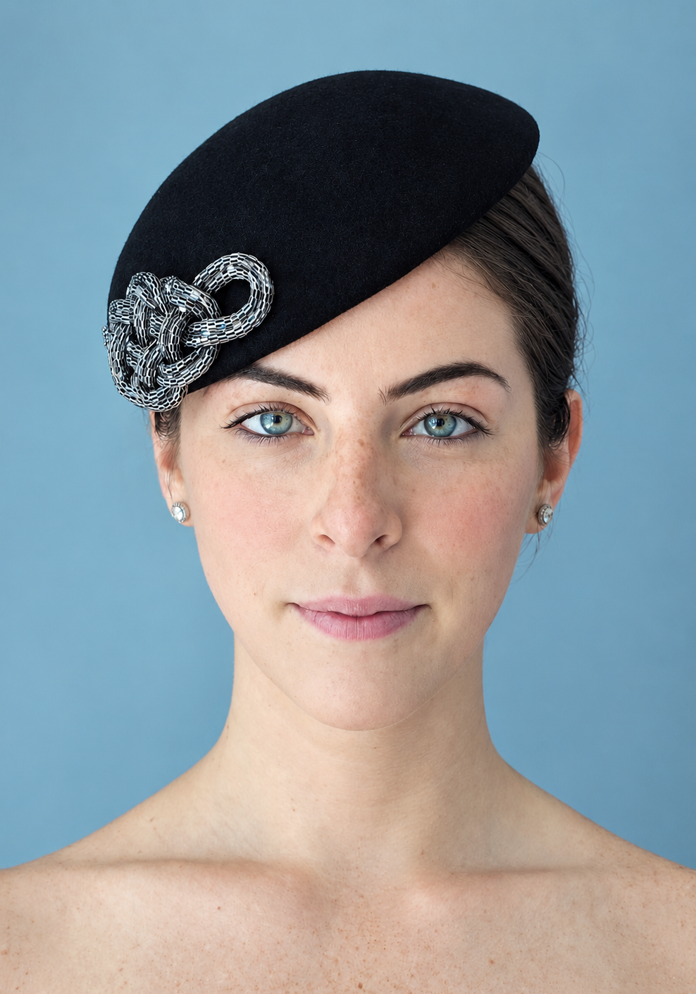 A woman wearing the Zoé noir felt percher hat with a silver chain-link decorative accent, photographed against a solid light blue background.