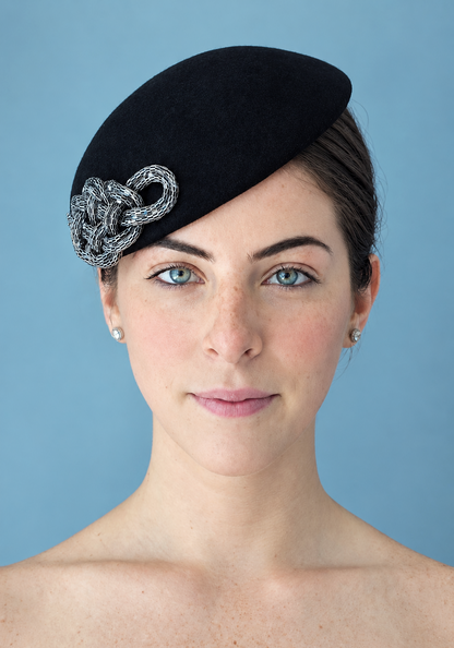 A woman wearing the Zoé noir felt percher hat with a silver chain-link decorative accent, photographed against a solid light blue background.